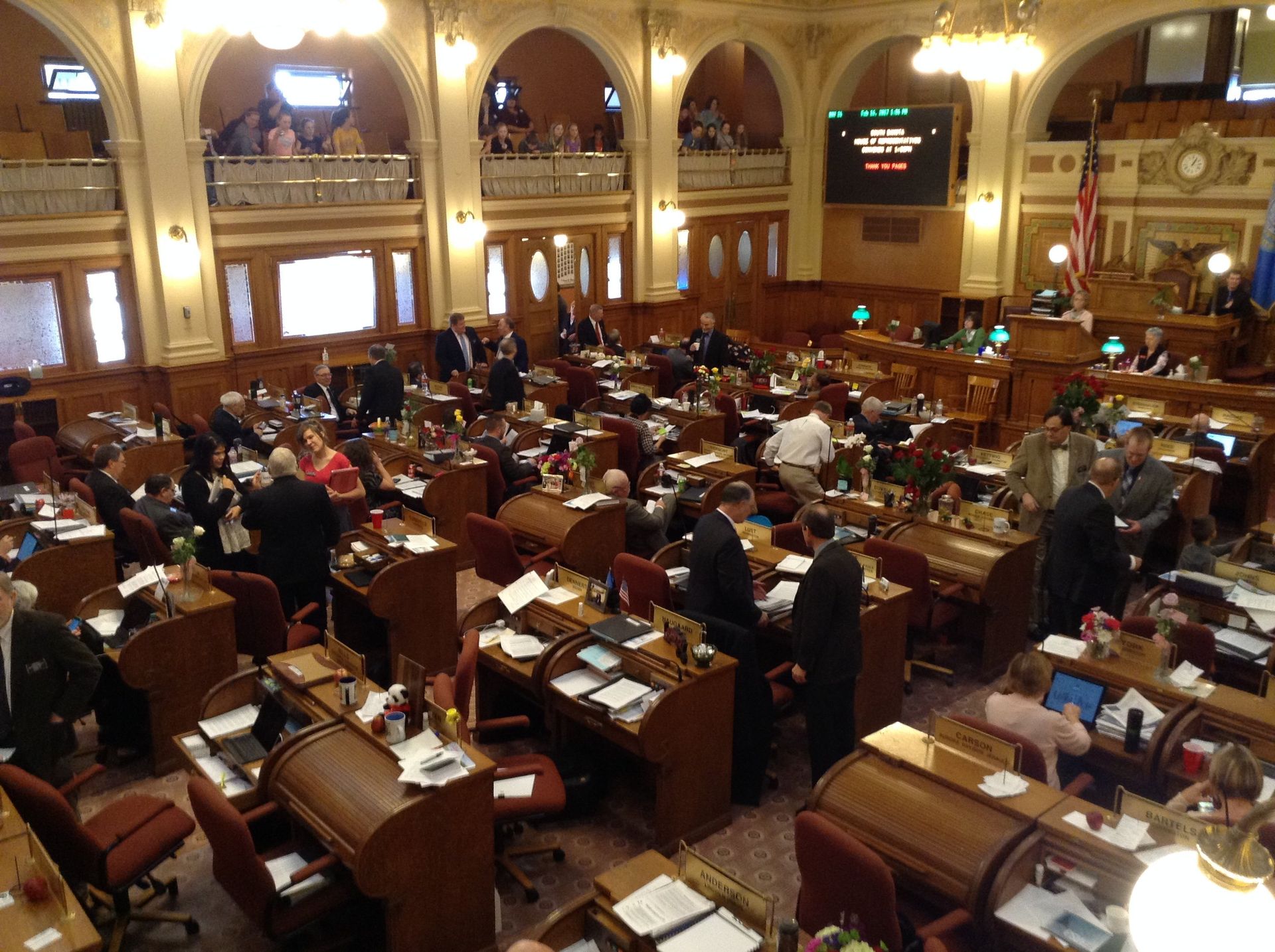 Interior of a legislative chamber with people seated at desks, some standing and talking.