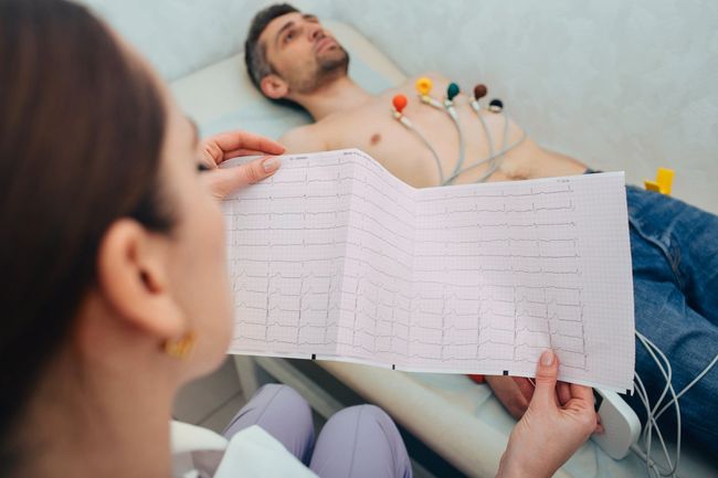 A healthcare professional examines an ECG readout while a patient lies on an exam table connected to a heart monitor.