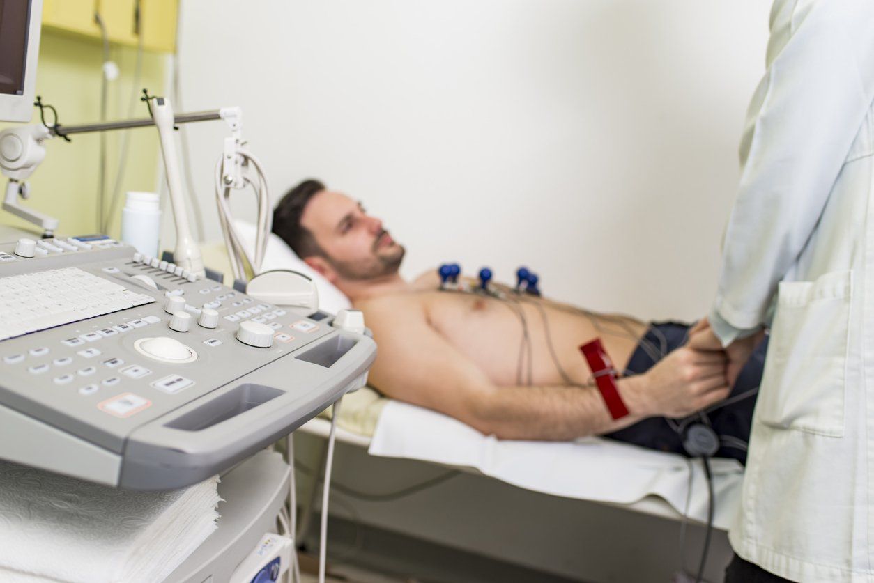 A healthcare provider monitors a patient lying on an exam table with ECG electrodes attached to his chest.