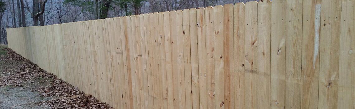 Wooden fence stretching along a tree-lined area. Boards are light brown. Ground is covered with leaves.
