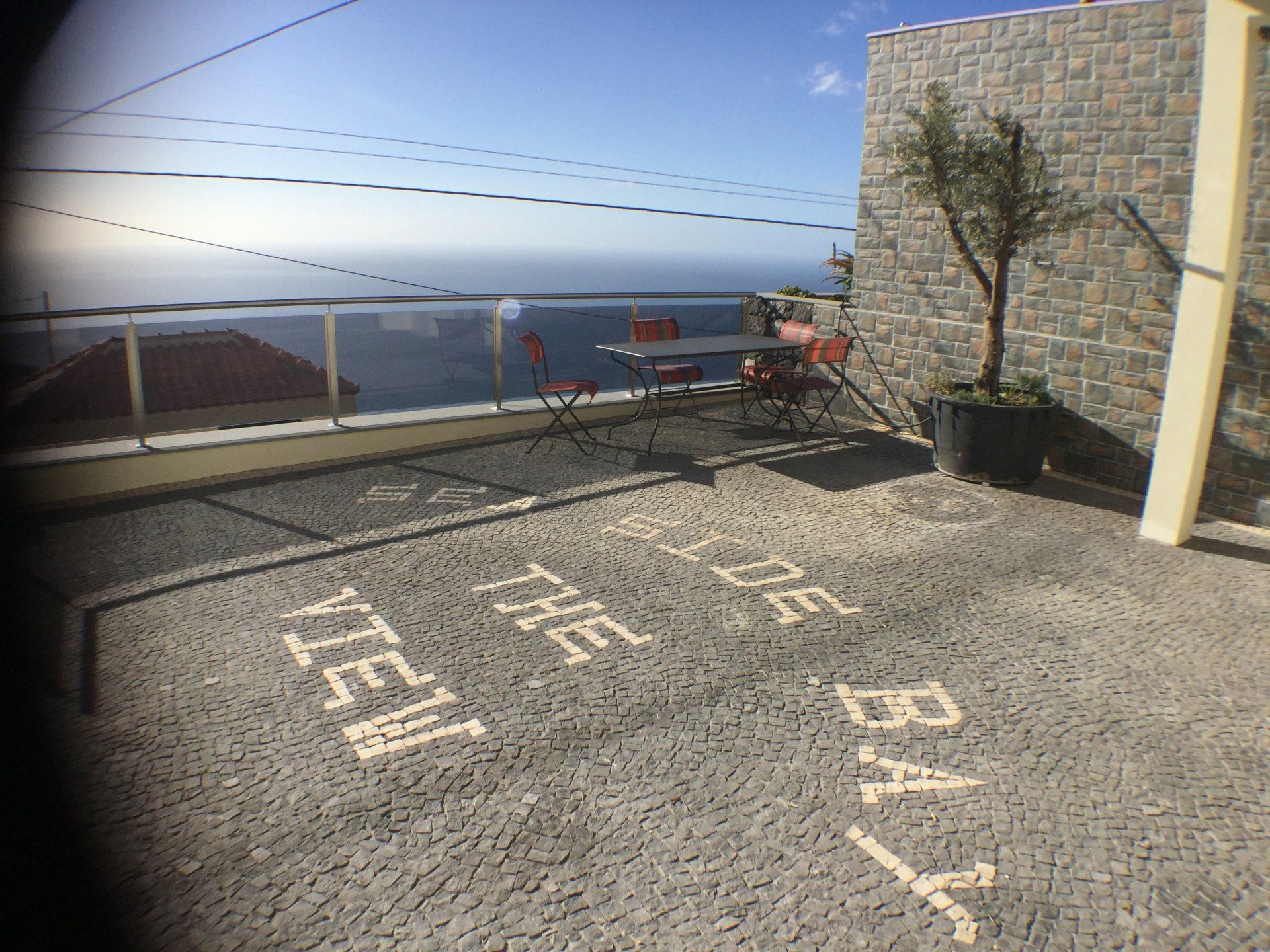 Terrasse mit Meerblick und dem gefliesten Schriftzug „THE VIEW OF THE BAY“. Tisch, Stühle, Topfbaum.