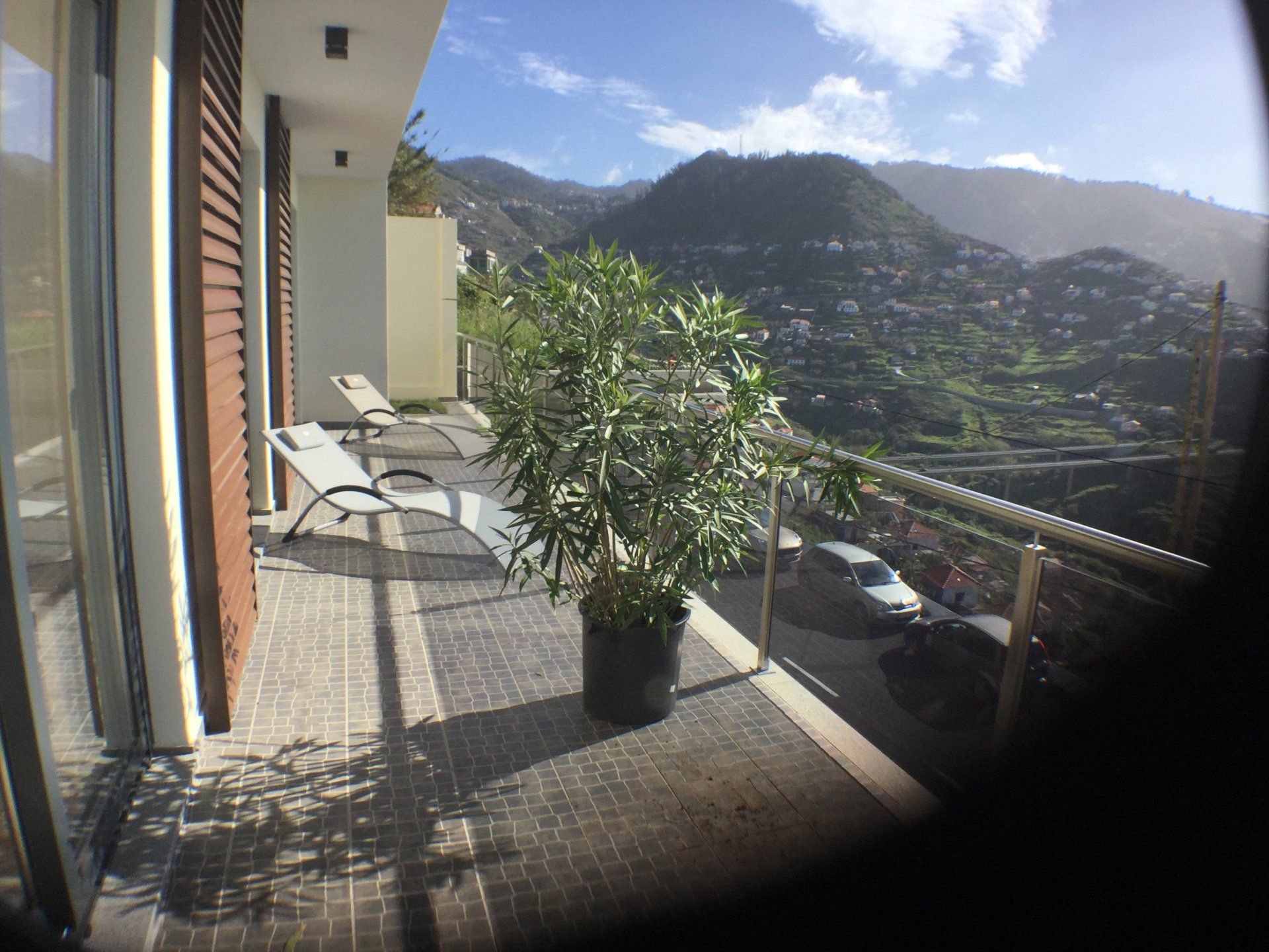Balkon mit Topfbaum, zwei Liegestühlen, Blick auf Berge und Häuser.