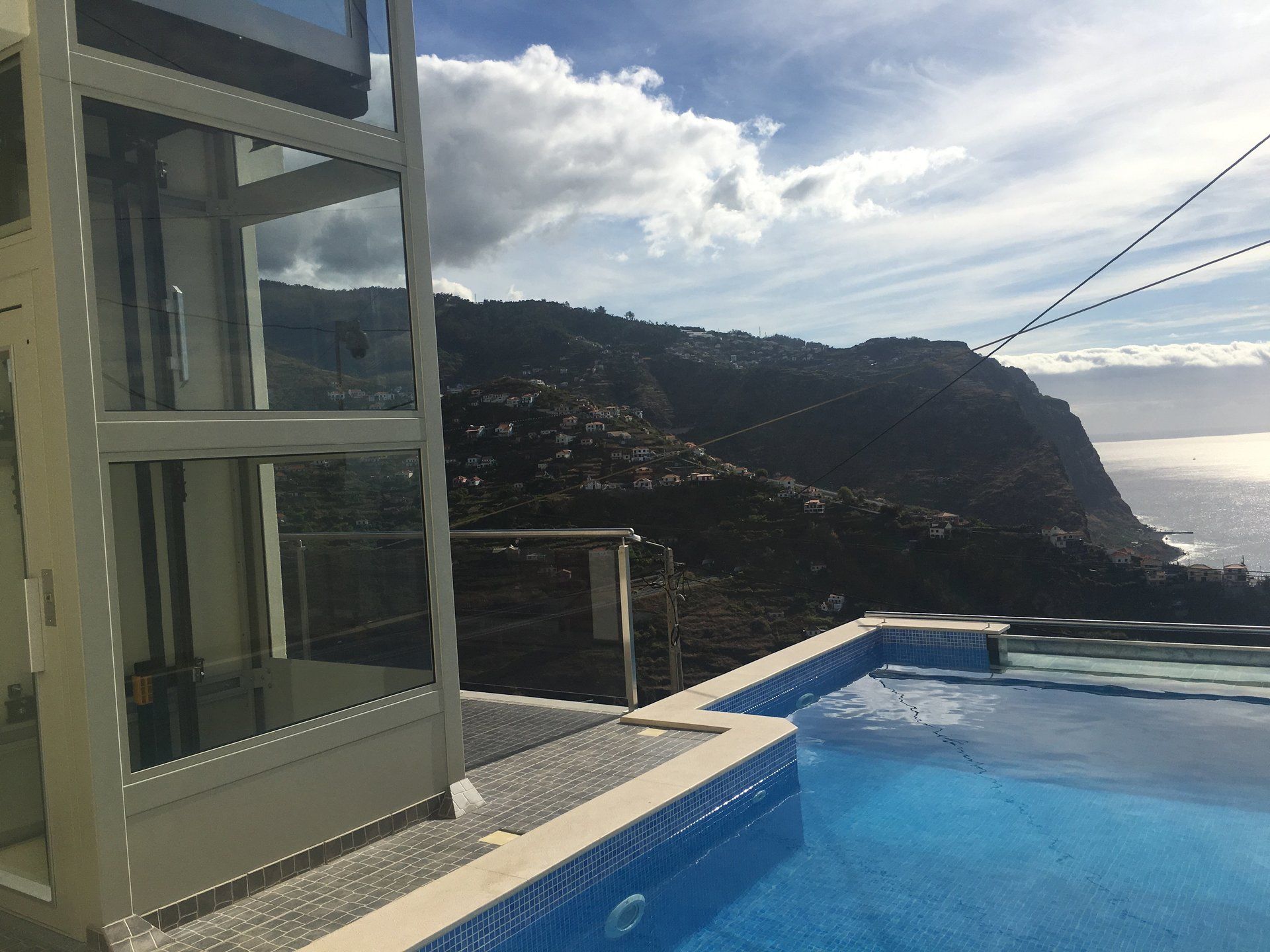 Ein gläserner Aufzug neben einem Pool mit Blick auf Berge und Meer. Sonniger Tag mit Wolken.