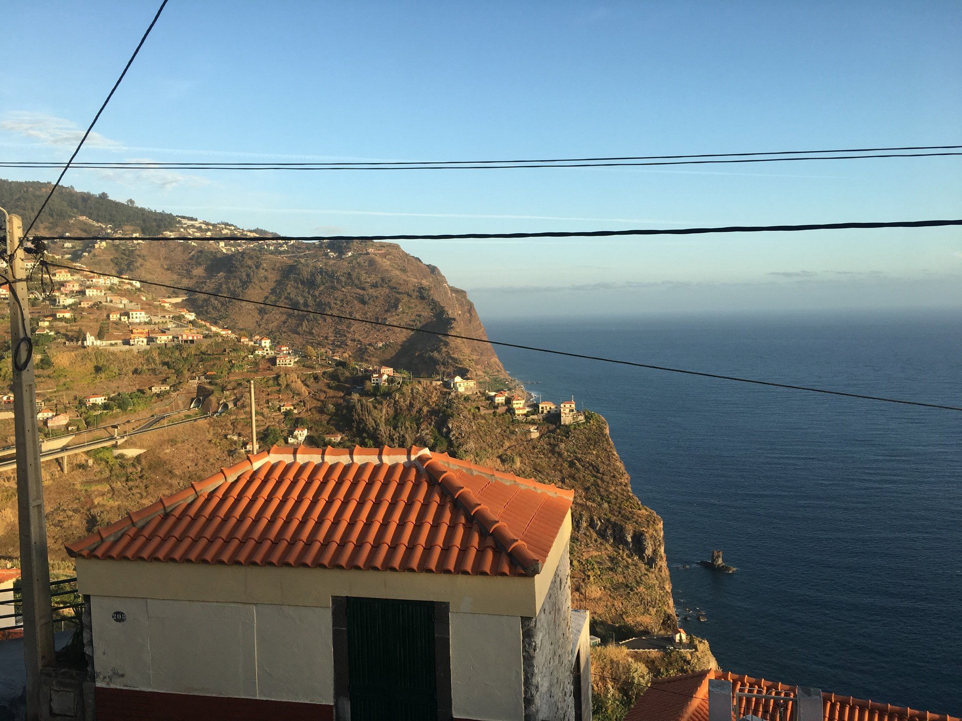 Ein Dorf am Hang mit Häusern und Meerblick unter blauem Himmel. Stromleitungen sind im Vordergrund zu sehen.