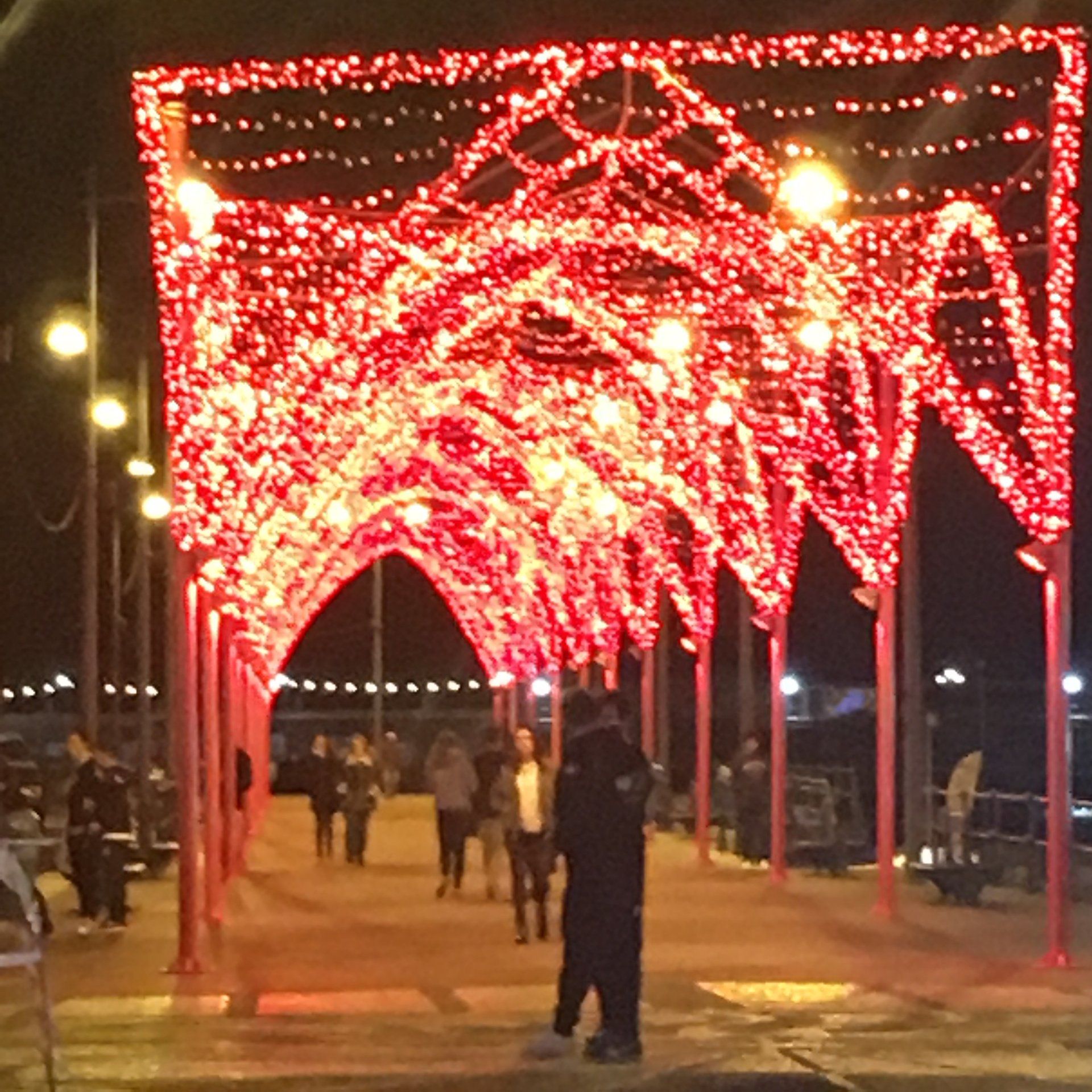 Rote Lichterketten bilden einen Bogen über einem Gehweg; die Leute gehen darunter hindurch.