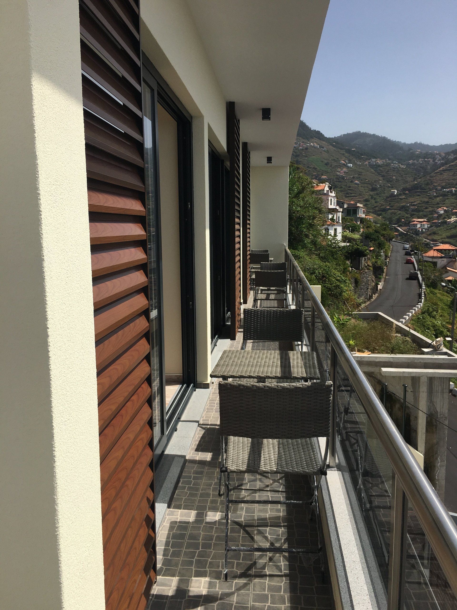 Balkon mit Korbmöbeln, Blick auf Straße und Berge. Braune Fensterläden, weiße Wände und klarer Himmel.