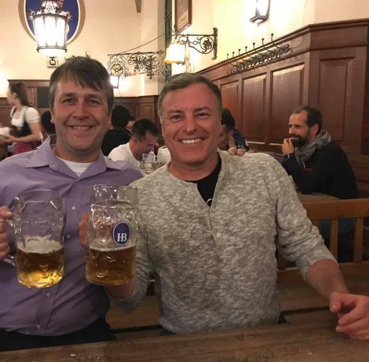 Two men holding beer steins, smiling in a Bavarian beer hall.