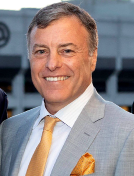 Man in light gray suit smiles, gold tie and pocket square, outside a building.