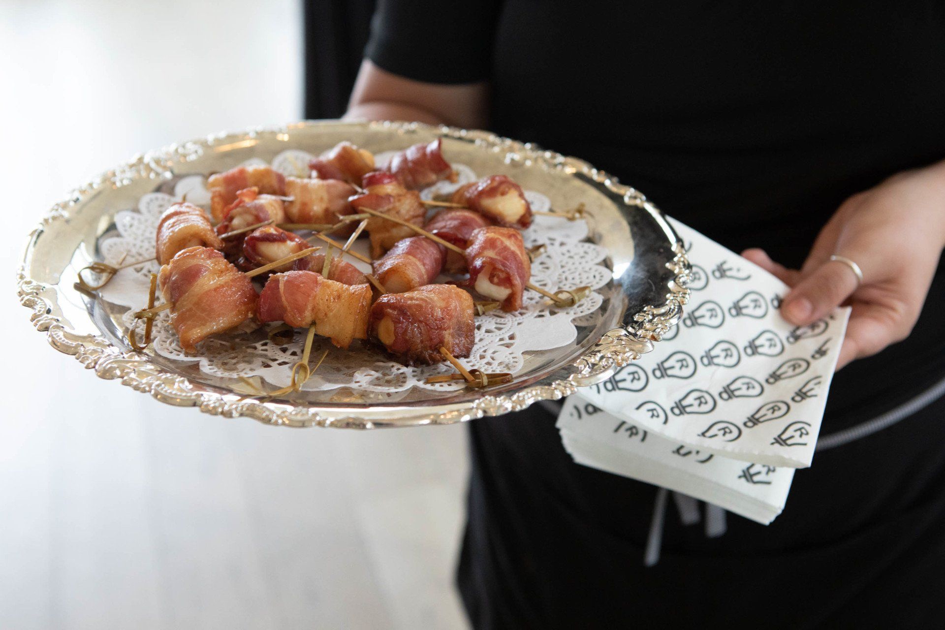 Person holding a silver platter of bacon-wrapped appetizers, accompanied by a stack of napkins.