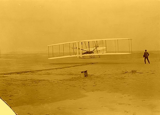 Wright brothers' first successful airplane in flight at Kitty Hawk, North Carolina; Orville in plane, man running alongside.