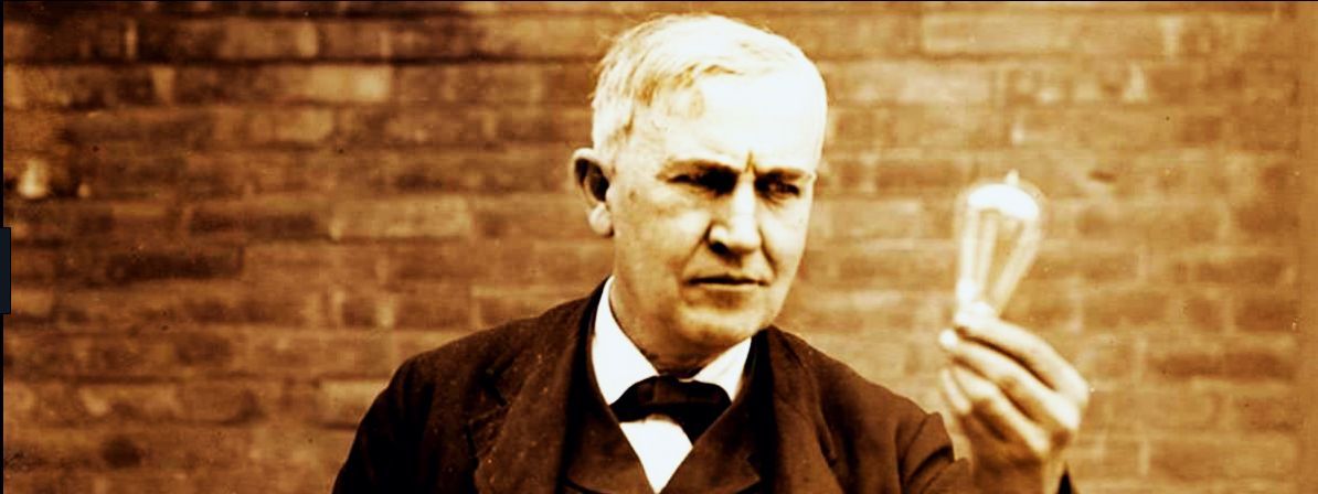 Thomas Edison holding a lit lightbulb in front of a brick wall, sepia toned.