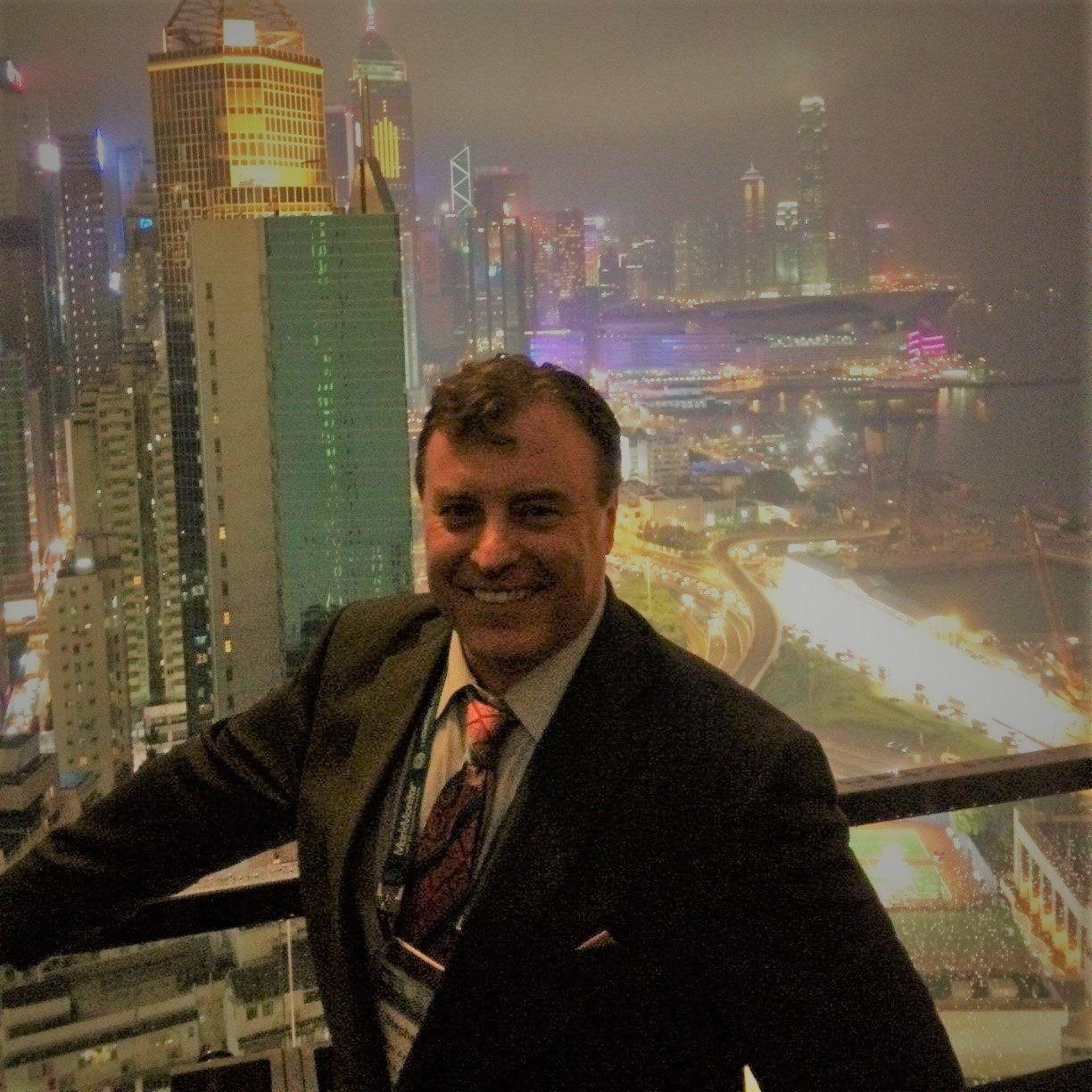 Man in suit smiles in front of a cityscape at night.
