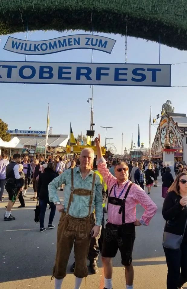 Two men in lederhosen pose at Oktoberfest under a sign reading 