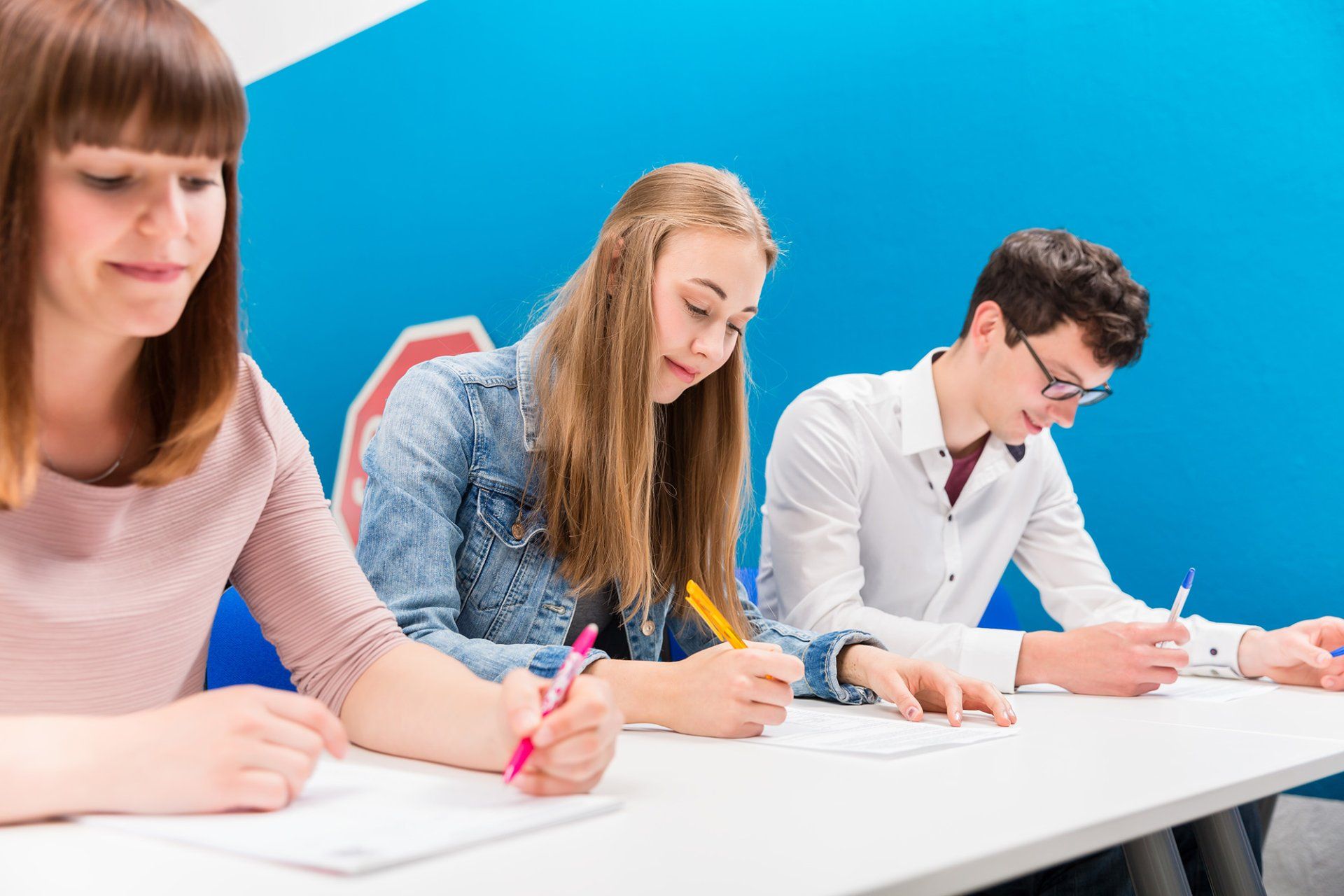 Tre studenti scrivono su un foglio di carta a una scrivania in un'aula luminosa.
