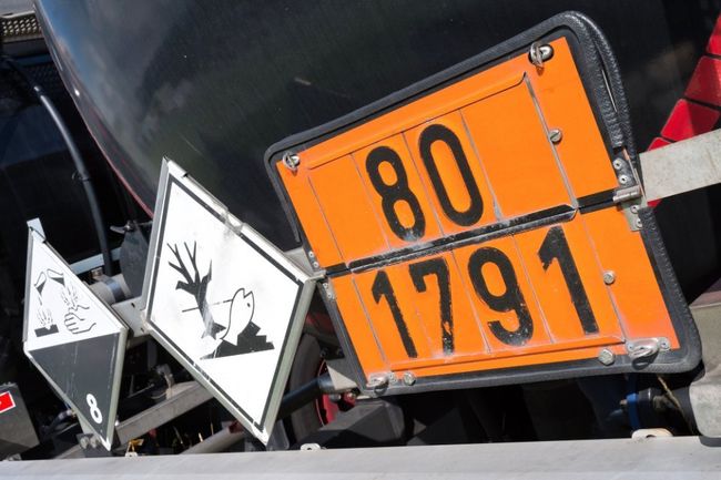 Cartelli arancioni di segnalazione di pericolo con i numeri 80 e 1791 su un veicolo, accanto a un segnale di avvertimento in bianco e nero.