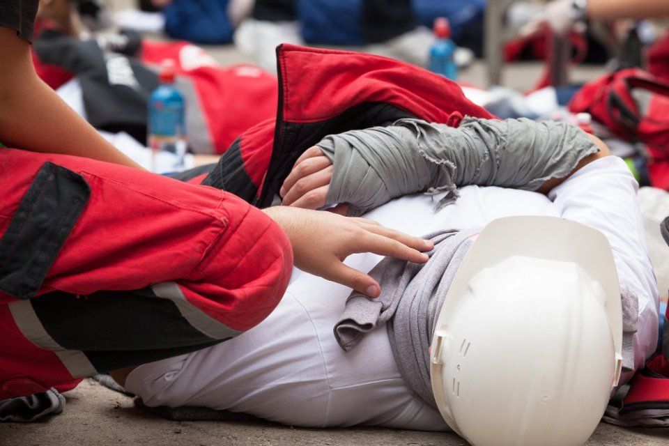 Durante un'esercitazione di emergenza all'aperto, una persona con il casco giace a terra mentre altre persone con giubbotti rossi le prestano soccorso.