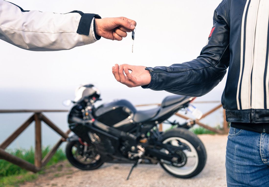 Due mani si scambiano le chiavi di una motocicletta davanti a una motocicletta nera parcheggiata all'aperto.
