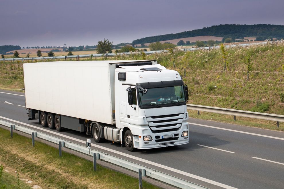 Un camion bianco percorre un'autostrada affiancato da un guardrail, con campi aperti sullo sfondo.