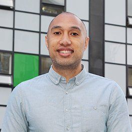 Man with a shaved head smiles in a light blue button-down shirt, in front of a modern building.