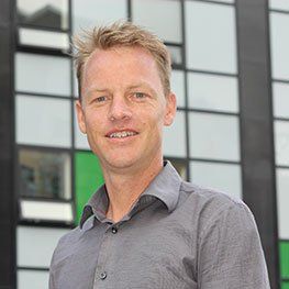 Man in gray shirt smiles, standing outside a building with green and glass windows.
