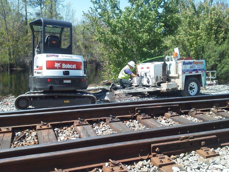 A Bobcat excavator and worker repairing railroad tracks. Sunny day, forest background.