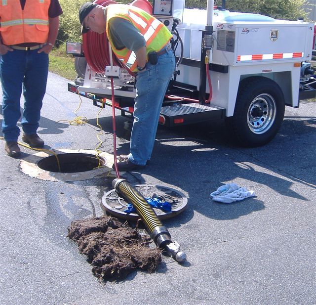 Two workers inspect a sewer, one looking down while equipment is used. Debris and open manhole are visible on the road.