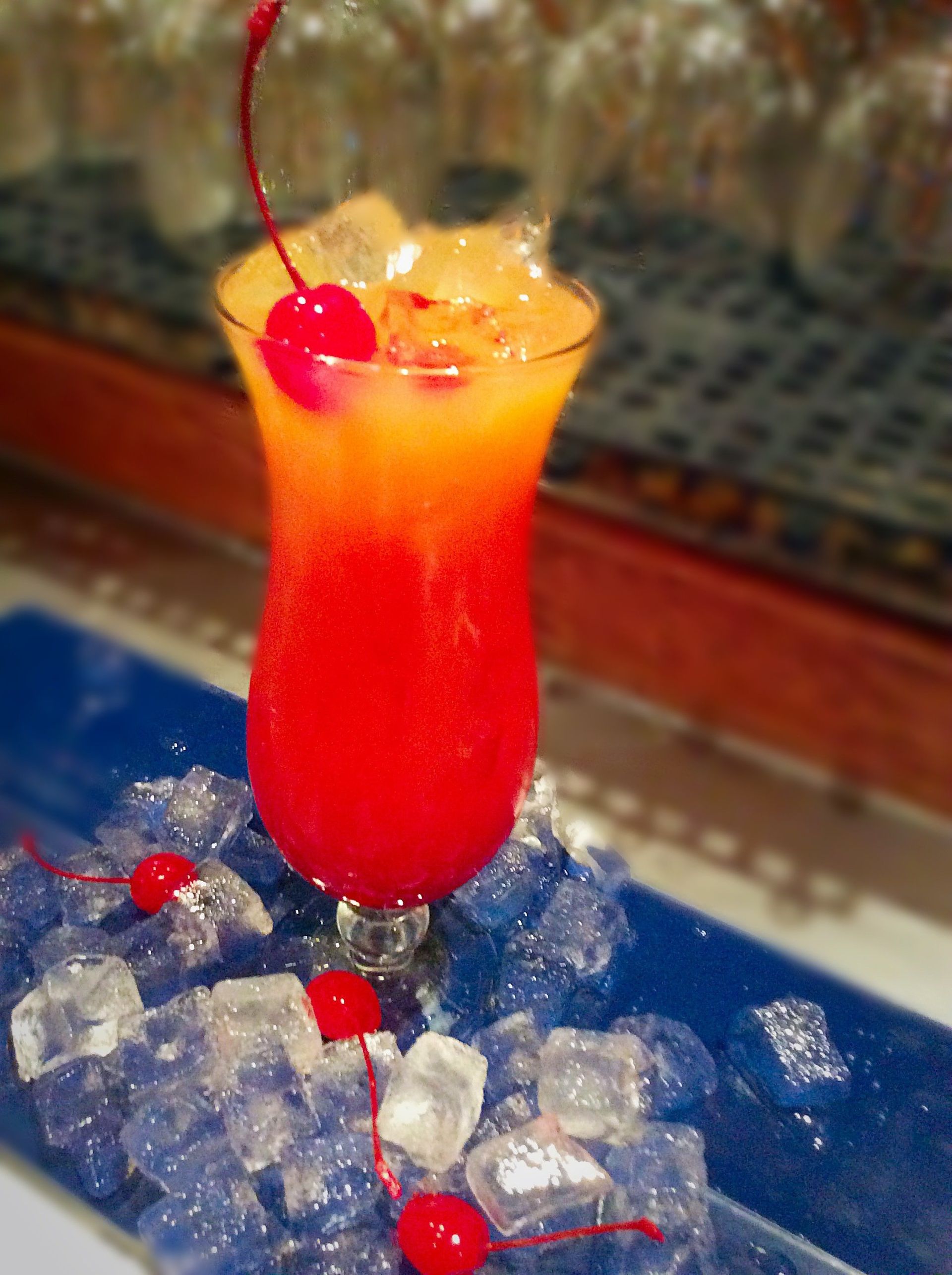 A layered cocktail in a tulip glass, red and orange, with a cherry garnish, surrounded by ice cubes.