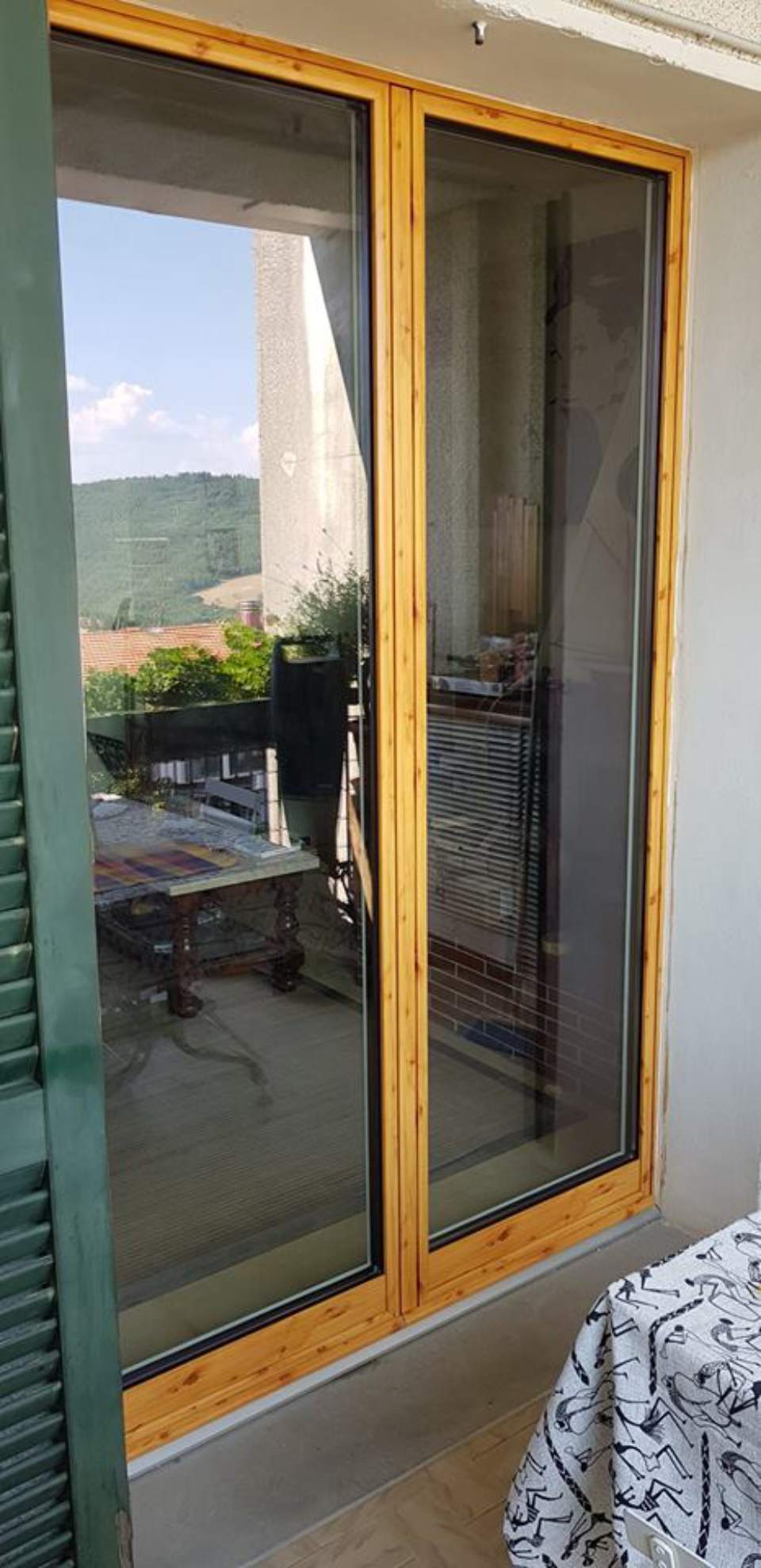 Porta scorrevole in vetro con telaio in legno e zanzariera, che riflette un patio esterno e le colline in lontananza.