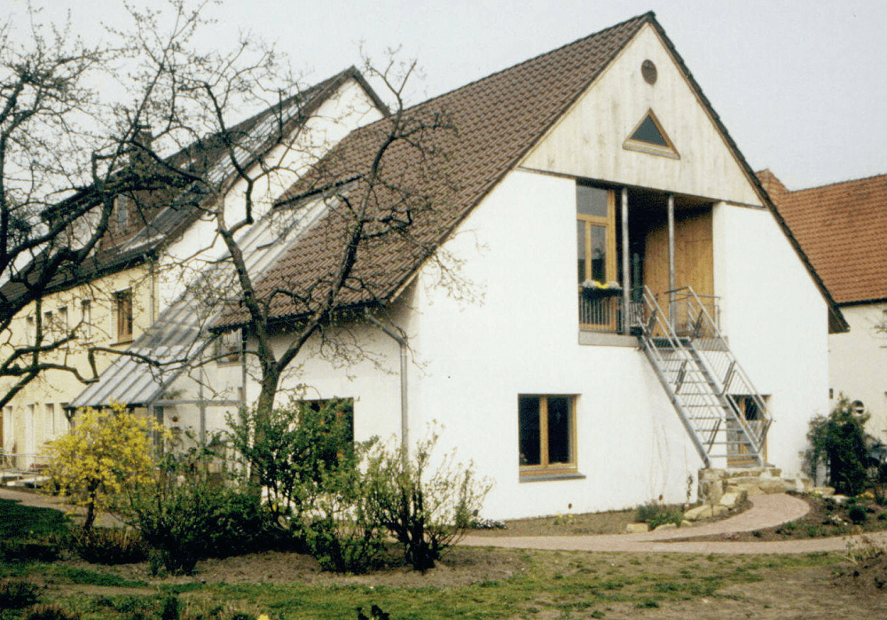 Weißes Gebäude mit braunem Dach und einer Außentreppe zum zweiten Stock. Im Vordergrund sind Bäume und Sträucher zu sehen.