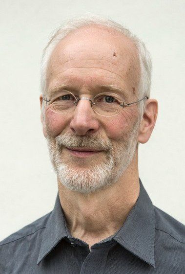 Ein Mann mit Brille, ergrauendem Haar und Bart, der ein graues Hemd trägt, lächelt in die Kamera.