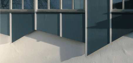 Blaue Gebäudefassade mit weißen Zierelementen, die Schatten auf die darunter liegende, strukturierte weiße Wand wirft.