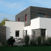 Modernes Haus mit weißen Wänden, dunklem Holz im oberen Bereich, roten Fensterrahmen und Außenterrasse.