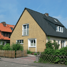 Ein gelbes Backsteinhaus mit schwarzem Dach, weiß gerahmten Fenstern und einem kleinen Holzzaun an einem sonnigen Tag.