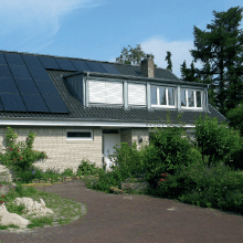 Haus mit Solarpaneelen auf dem Dach, Dachgauben und üppigem Grün davor.