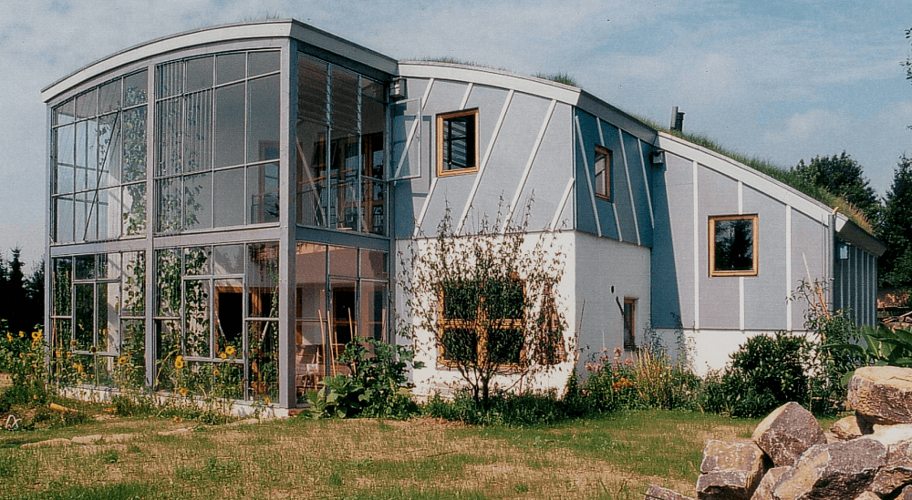 Modernes, zweistöckiges Haus mit geschwungenem Glaswintergarten, hellblauer Fassade, Gründach und naturbelassenem Vorgarten.