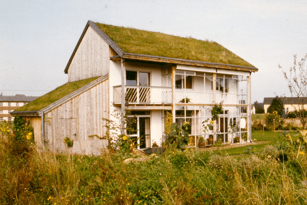Zweistöckiges Haus mit Gründach, Holzverkleidung und verglastem Balkon, umgeben von hohem Gras.