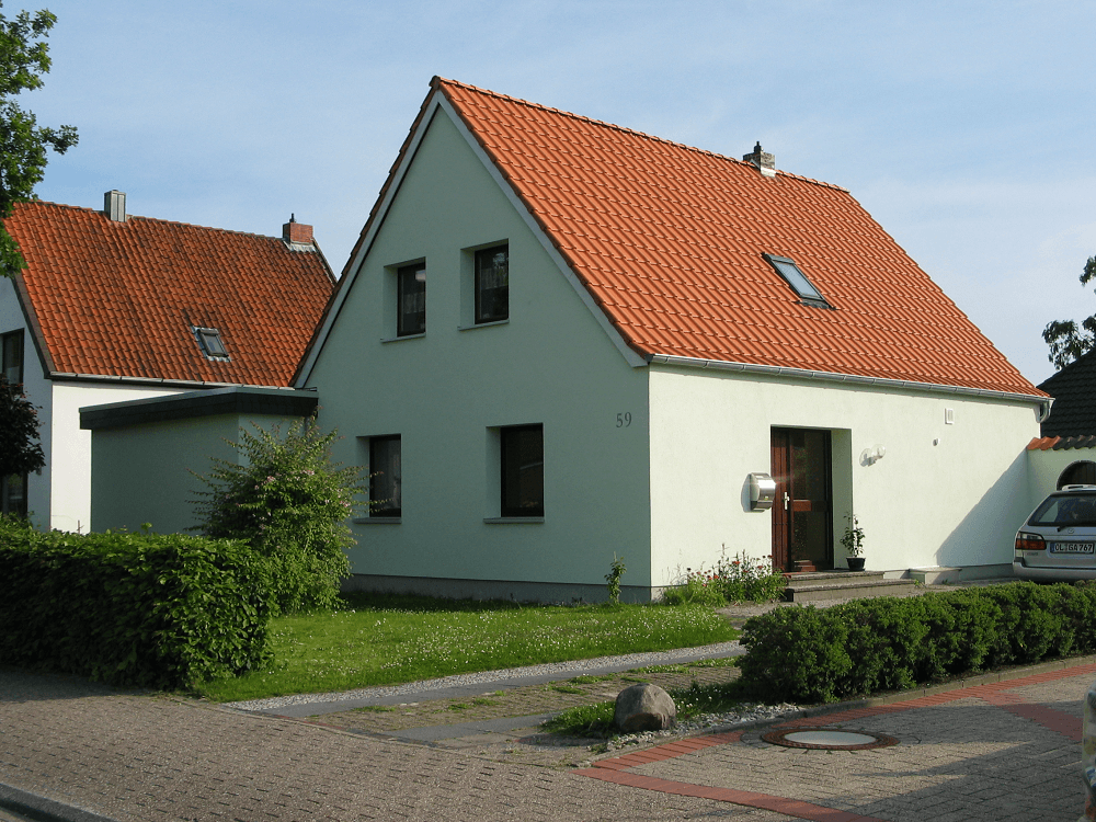 Hellgrünes Haus mit orangefarbenem Ziegeldach, mehreren Fenstern und kleinem Vorgarten mit Büschen.