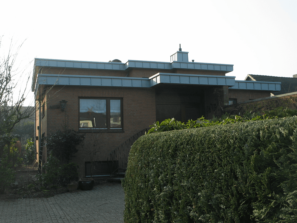 Backsteinhaus mit Flachdach und Metallverkleidung, umgeben von Grün und einer Auffahrt.