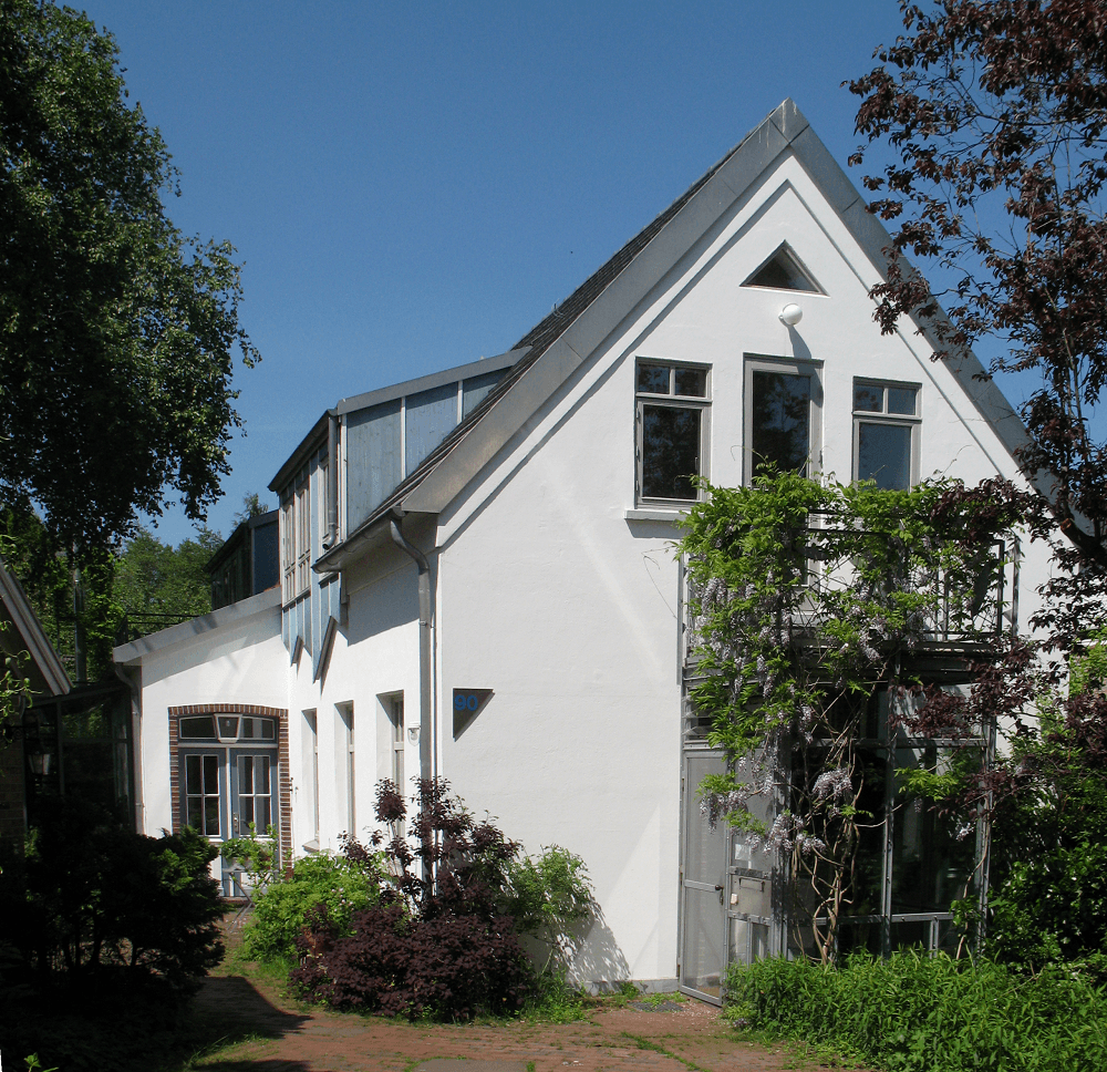 Weißes Haus mit Giebeldach und grauen Metallverzierungen, umgeben von Grün.