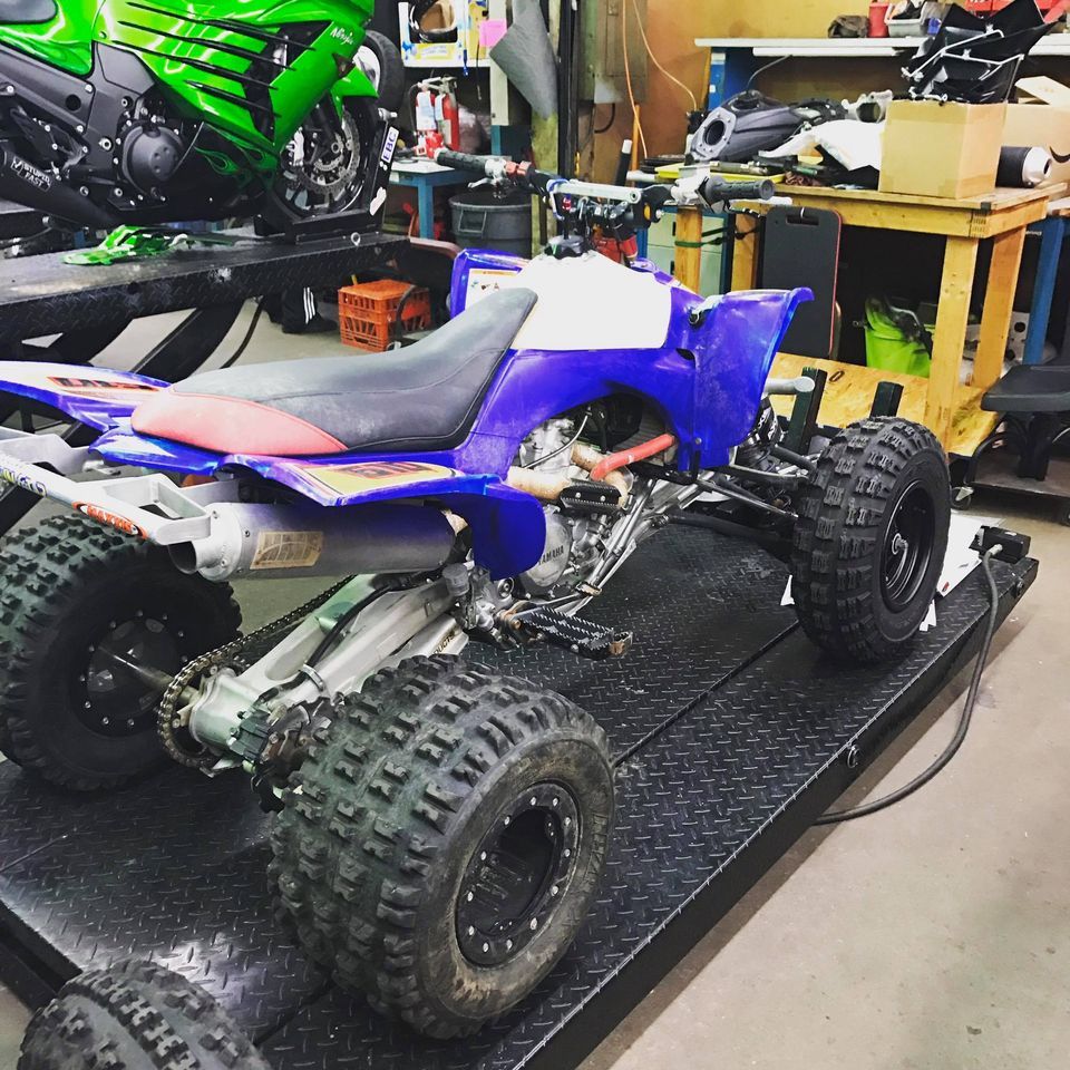 A blue and white ATV sits on a metal lift in a workshop, with a green motorcycle visible in the background.