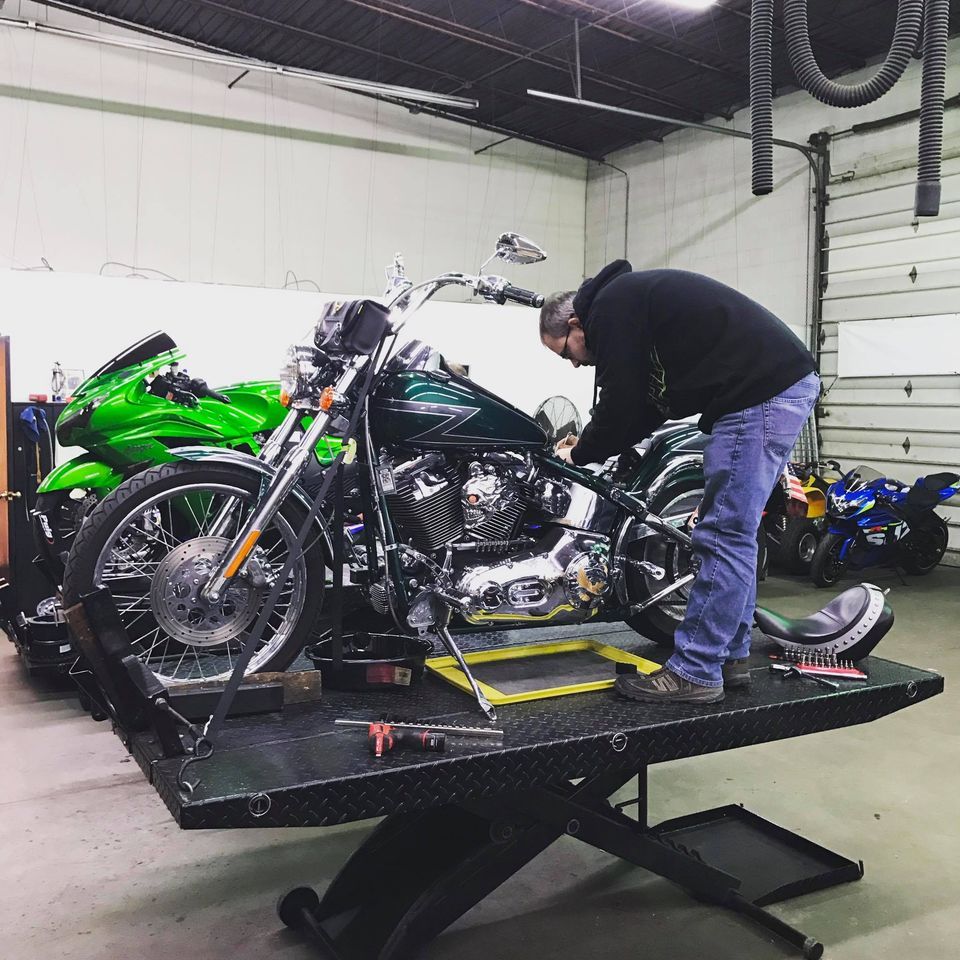 A mechanic in a garage works on a dark green cruiser motorcycle mounted on a hydraulic lift, with other bikes nearby.