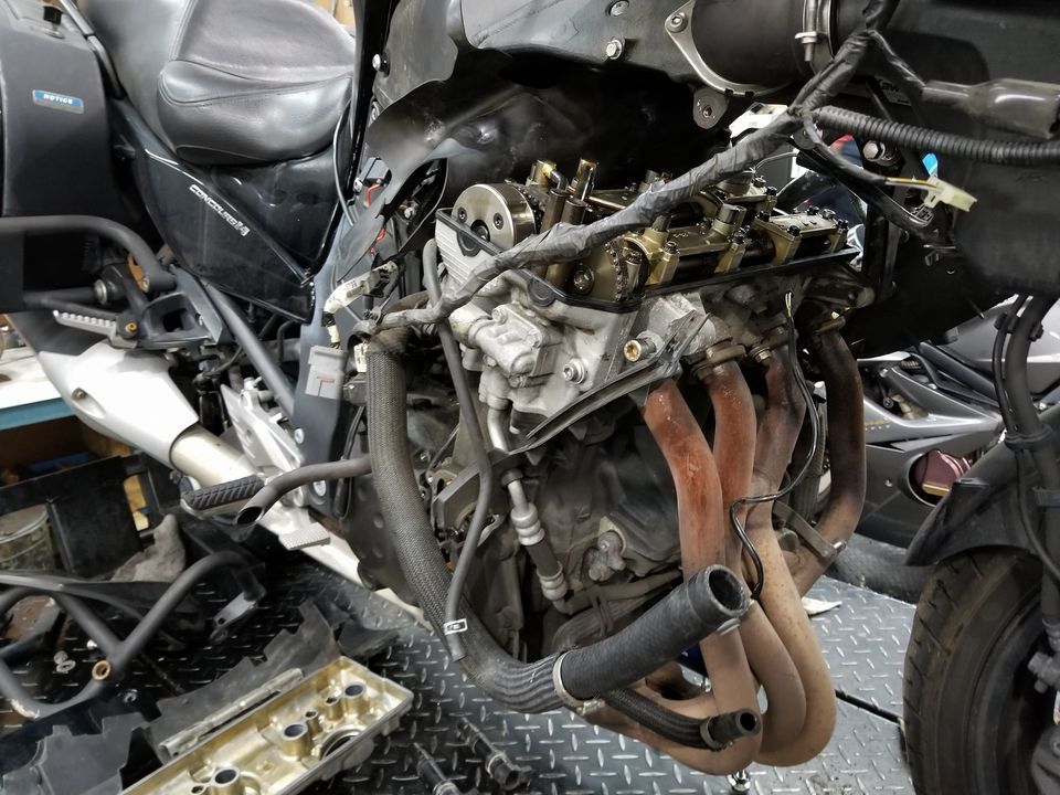 A disassembled motorcycle engine with exposed valve train, header pipes, and removed cover in a repair shop.