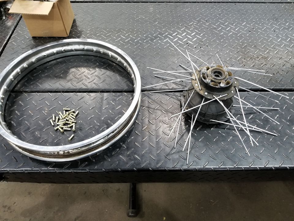 A disassembled motorcycle wheel with spokes attached to the hub, a metal rim, and a small pile of nipples on a shop table.