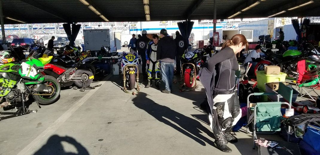 A group of people prepare motorcycles under a large outdoor covered structure at a track day or racing event.