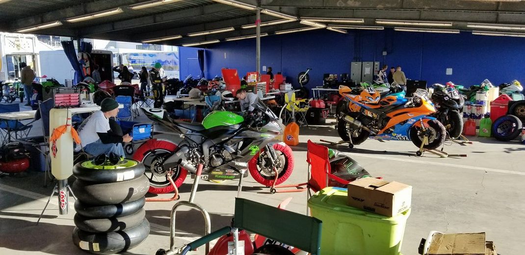 A pit lane area with racing motorcycles parked under a covered structure, with crew members and equipment in the background.
