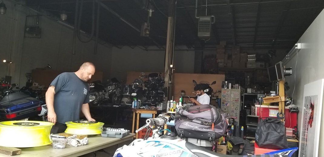 Two people work on motorcycle parts inside a cluttered repair shop with various tools and equipment visible.