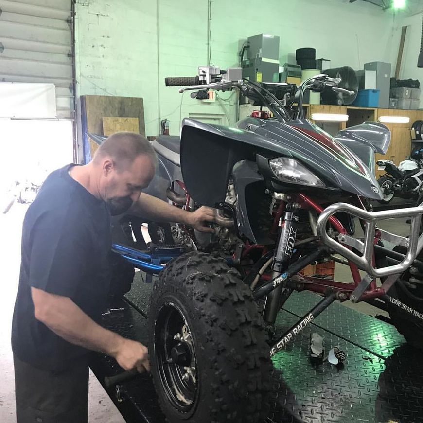 A person works on a grey all-terrain vehicle in a garage, focusing on the front wheel and suspension.