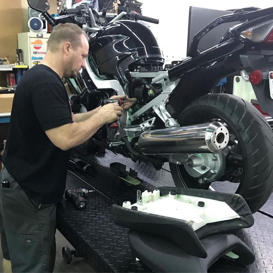 A technician repairs a black motorcycle on a lift in a workshop, with the bike's seat removed and set aside.