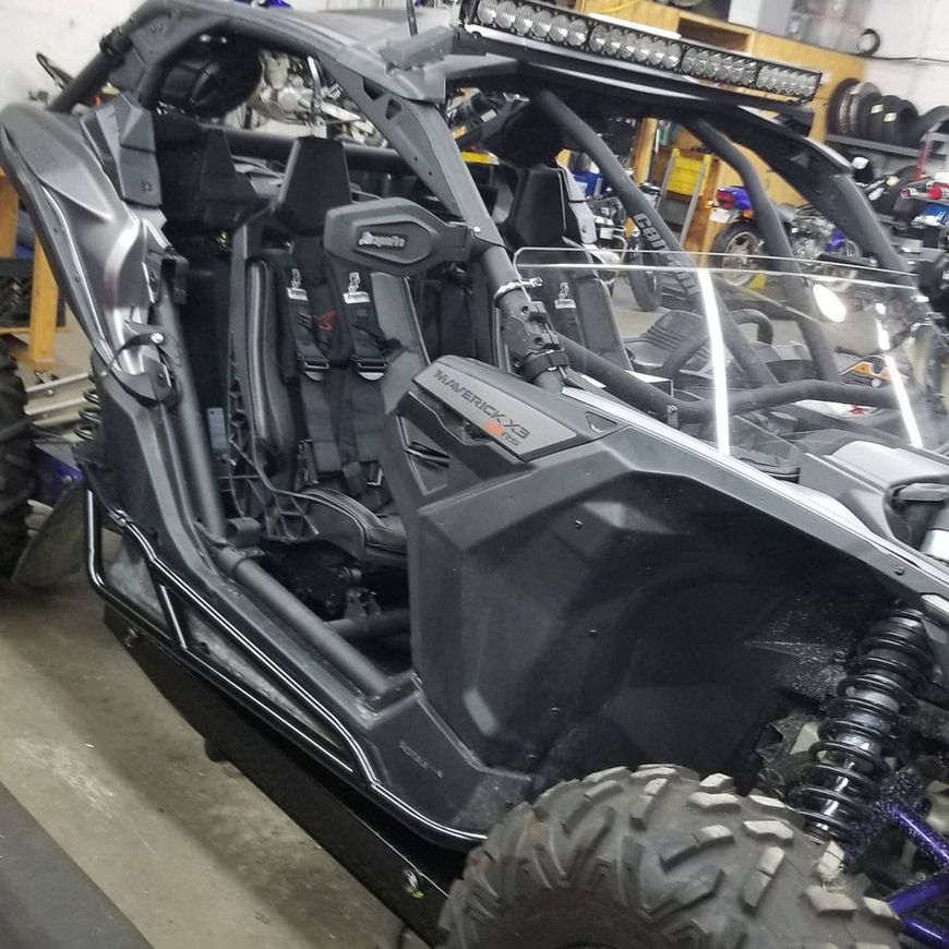 A gray Can-Am Maverick X3 off-road vehicle with a roof-mounted LED light bar parked inside a workshop.