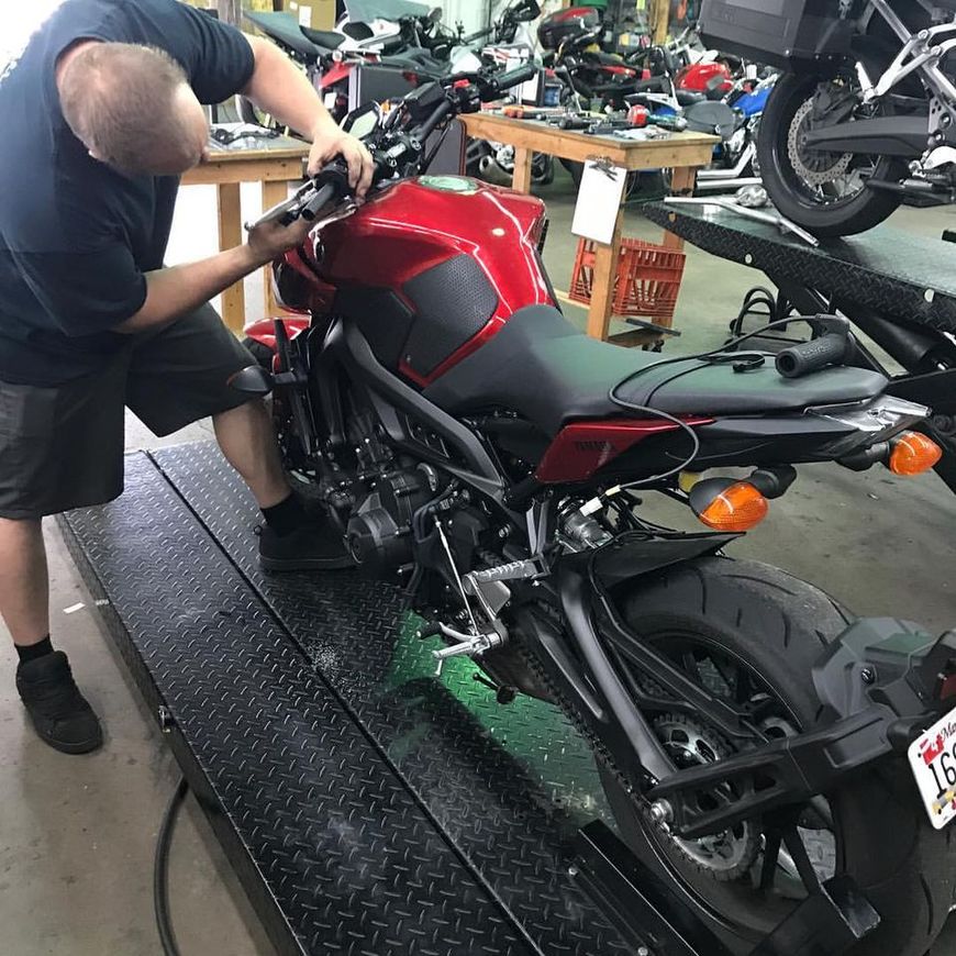A mechanic works on the handlebars of a bright red motorcycle parked on a lift inside a repair shop.